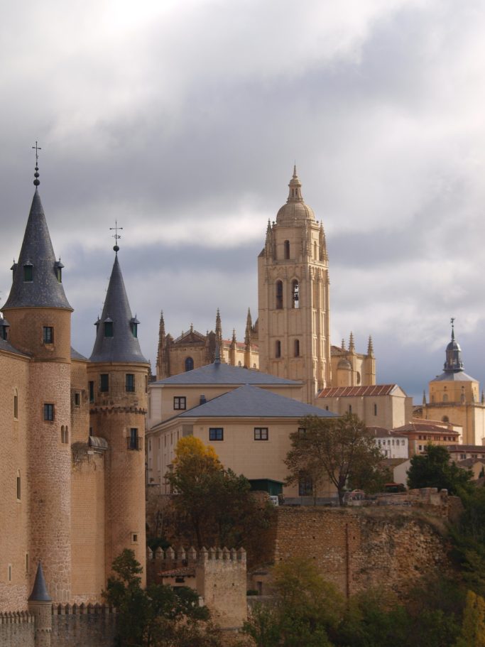 Castillo con torres y catedral al fondo, rodeados de nubes grises.