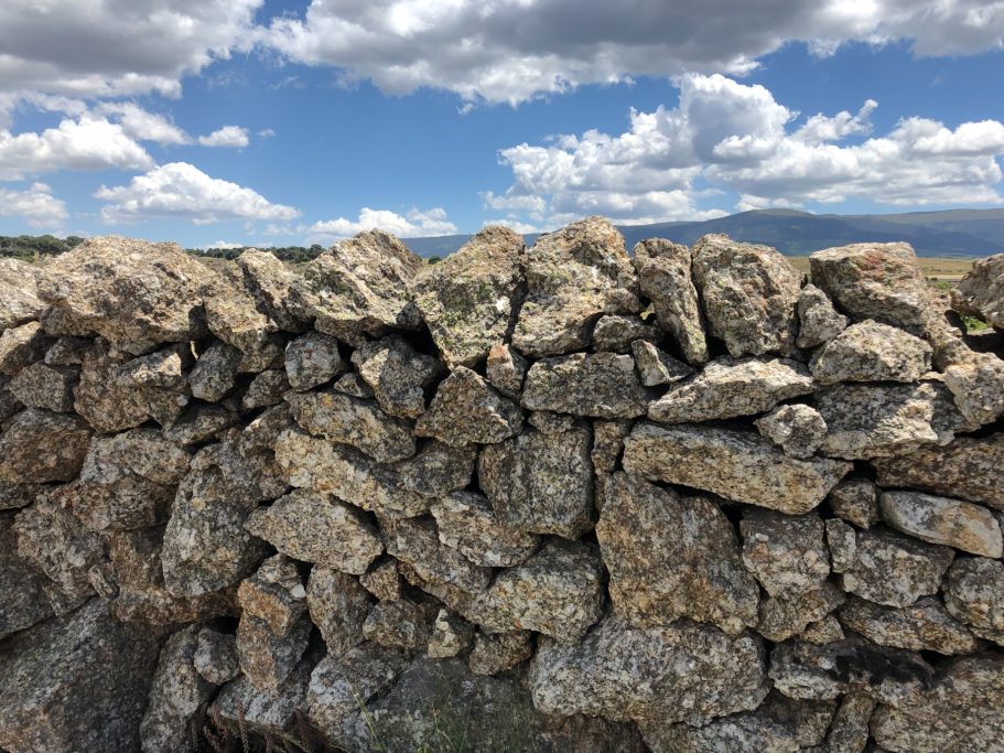 Pared de piedras apiladas bajo un cielo nublado con montañas al fondo.