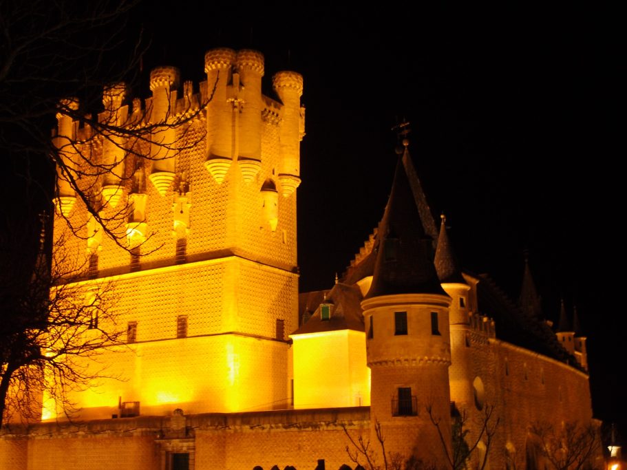 Castillo iluminado en la noche, con torres y murallas resaltadas en amarillo.