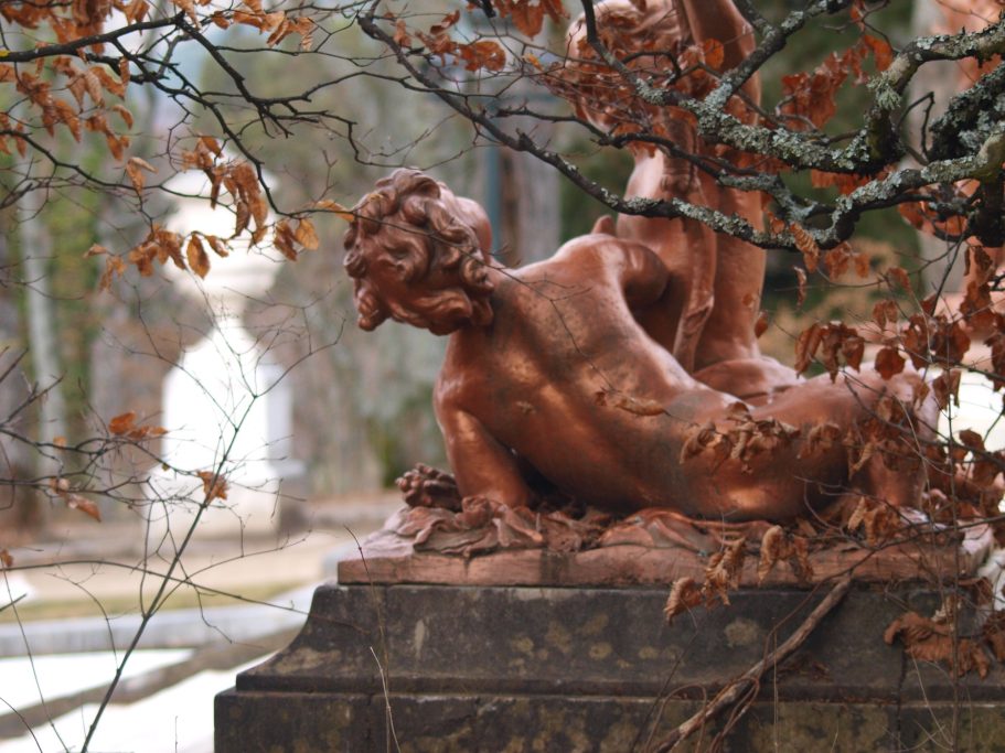Escultura de un niño con forma de pez, rodeada de hojas secas y un fondo de jardín.