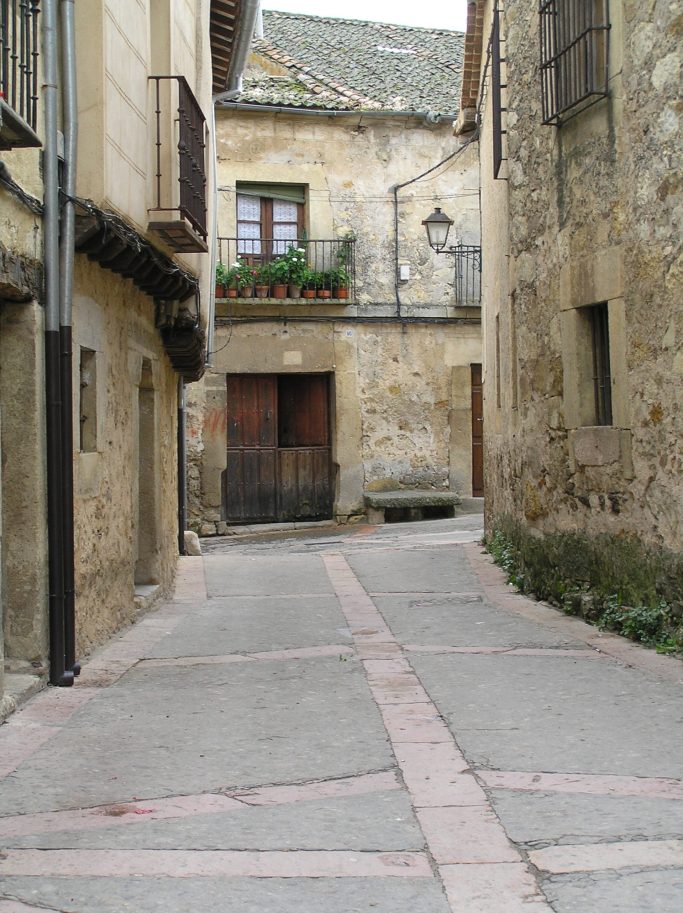 Callejón empedrado con casas de piedra y un balcón con plantas.
